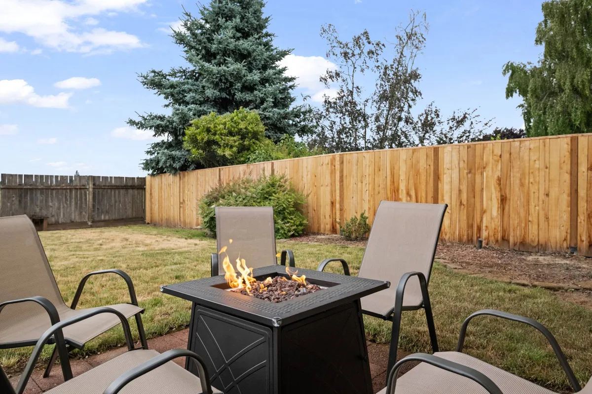A cozy firepit area with lawn chairs arranged in a circle, bordered by a wooden fence and mature trees. The late afternoon light emphasizes a tranquil, inviting atmosphere for gatherings around the fire as the sun sets.