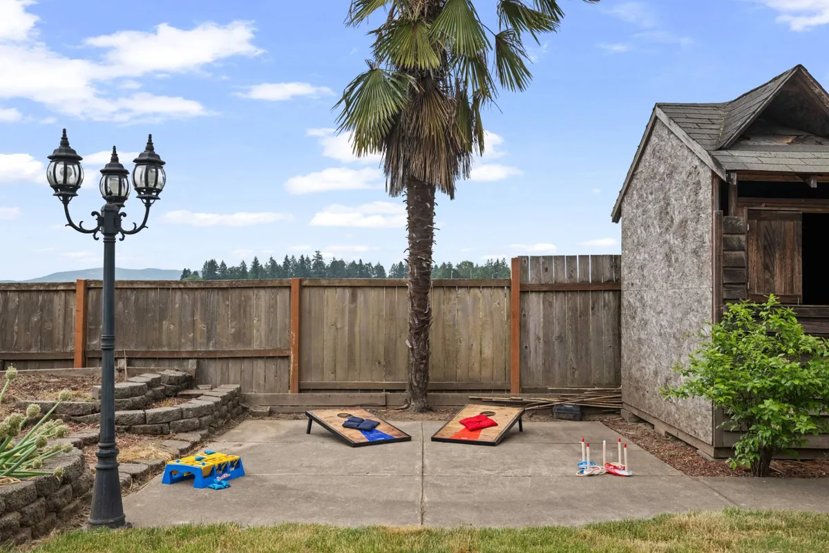 The exterior shot captures a palm tree shading a social space with outdoor games set up, a lantern-style lamp for evening ambiance, and an old shed in the background. The area invites friendly competition or relaxed evenings in a casual, pet-appropriate environment.