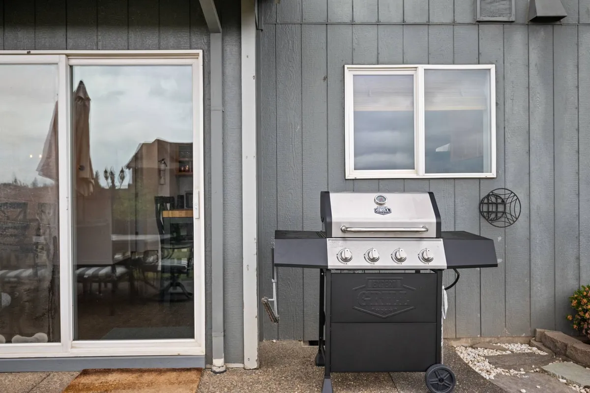 The photo showcases an exterior patio area with a grill, a large window looking into the house, and ample outdoor seating. The space looks inviting for gatherings, dining al fresco, or simply relaxing while keeping an eye on pets as they play nearby.