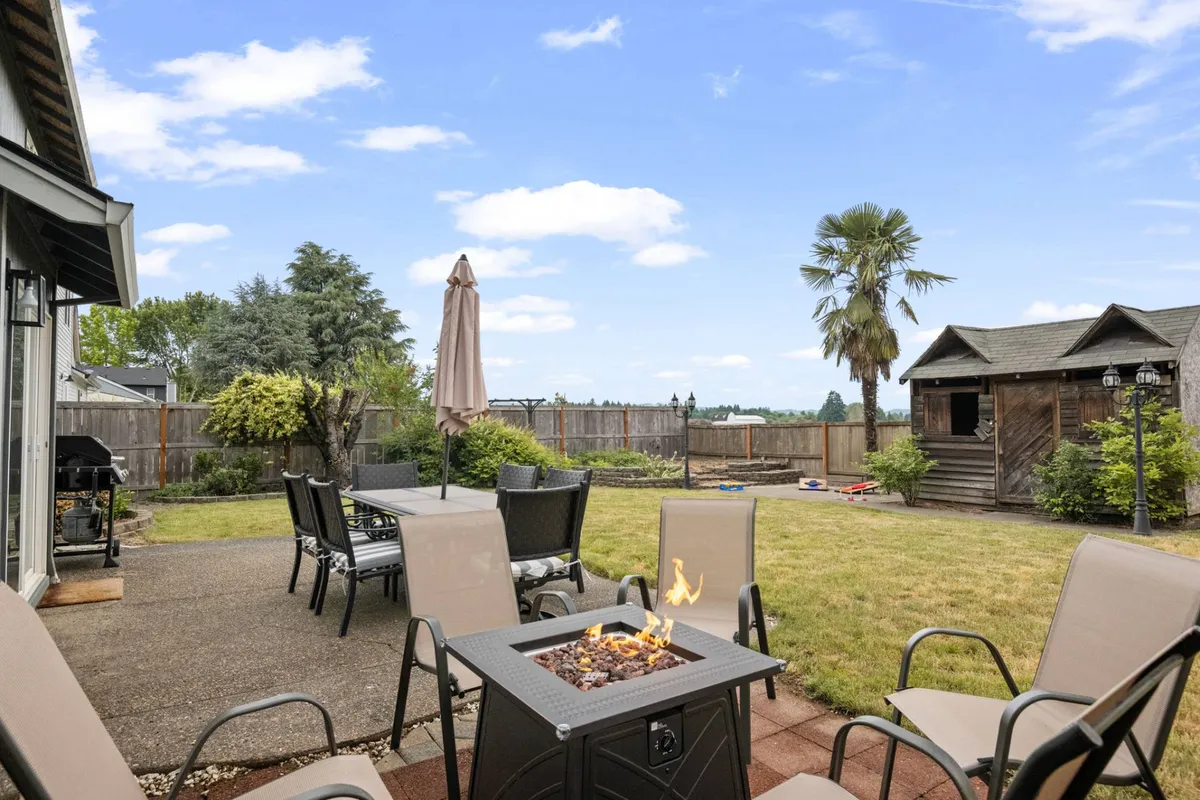 A cozy outdoor patio featuring comfortable seating around a fire pit, surrounded by lush greenery and a palm tree. The space exudes a relaxed and social mood, perfect for unwinding with a glass of wine after a day of tastings.
