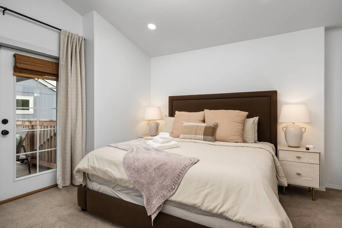 A serene bedroom featuring a plush bed adorned with soft pillows and throws, nightstands with warm lamps, and a sliding door that leads directly out to a private deck. Natural light fills the space, highlighting the tranquil design and the inviting transition to the outdoor balcony area.