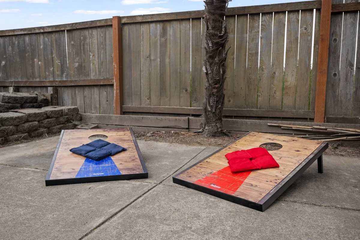 This image features a backyard scene with cornhole boards set up on the lawn, colorful bags ready for play, and a sturdy wooden fence enclosing the area. The overall atmosphere is playful and relaxed, with space for both social games and outdoor relaxation. The securely fenced yard is clearly visible, reinforcing the pet-friendly aspect.